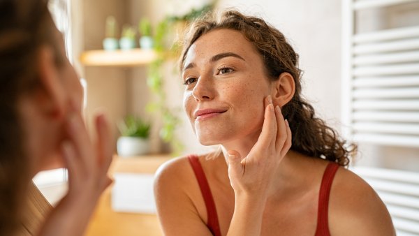Beauty girl looking at mirror while touching her face and checking pimple, wrinkles and bags under the eyes, during morning beauty routine. Smiling pretty woman applying cream on her face in bathroom. Happy smiling beautiful young woman applying moisturizer.