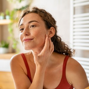Beauty girl looking at mirror while touching her face and checking pimple, wrinkles and bags under the eyes, during morning beauty routine. Smiling pretty woman applying cream on her face in bathroom. Happy smiling beautiful young woman applying moisturizer.