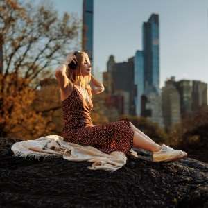 A woman sits serenely with a blanket, gazing at a city skyline from a vantage point, invoking a sense of peace despite the busy city backdrop