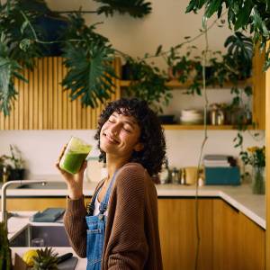 Happy Woman Enjoying A Fresh Green Smoothie In A Sunny Kitchen Decorated With Indoor Plants
