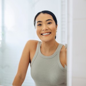 Cheerful Woman standing at her sink in the bathroom, looking at herself in the mirror.
