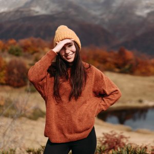 Cute young woman in a sweater and hat in the mountains by the lake in the autumn season.