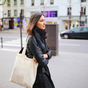 Vintage toned image of a young Parisian woman walking the city center, near Montmartre, at Boulevard de Clichy. She is wearing a black leather jacket and a scarf, carrying a tote bag. Shot with wide angle large aperture prime lens for more shallow depth of field in motion.