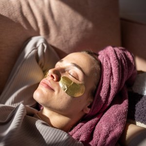 Young woman with eyes closed relaxing on sofa with towel wrapped around hair and gel patches under eyes.
