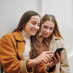 Two smiling young female friends swiping through an app on a smart phone while standing together on a city sidewalk