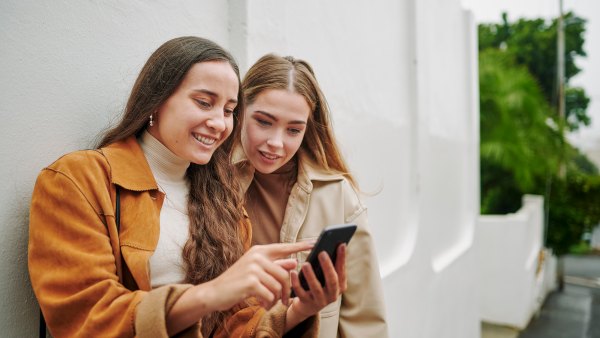 Two smiling young female friends swiping through an app on a smart phone while standing together on a city sidewalk