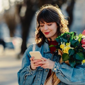 a woman holding a phone with a bouquet of flowers in a tree lined street