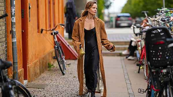 A guest wears silver earrings, a silver necklace, a black V-neck silk / satin long skirt, a dark brown suede long coat, black shiny varnished leather laces loafers, a pale yellow khaki shiny leather handbag , outside Remain, during the Copenhagen Fashion Week Spring/Summer 2024 on August 08, 2023 in Copenhagen, Denmark.