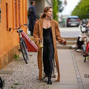 A guest wears silver earrings, a silver necklace, a black V-neck silk / satin long skirt, a dark brown suede long coat, black shiny varnished leather laces loafers, a pale yellow khaki shiny leather handbag , outside Remain, during the Copenhagen Fashion Week Spring/Summer 2024 on August 08, 2023 in Copenhagen, Denmark.