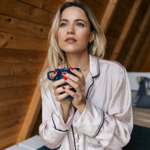 Beautiful young woman relaxing and feeling carefree in mountain house while drinking coffee or tea. She is sitting on bed in the bedroom
