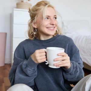 Lifestyle portrait of young woman sitting on bedroom floor with cup of tea, drinking from big white mug and looking aside, smiling happily.