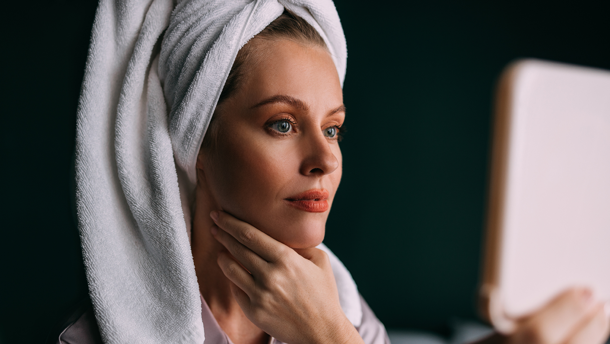 A smiling Caucasian female with her hair wrapped up in a towel keeping her face clean and hydrated using different cosmetic products. (skincare concept)