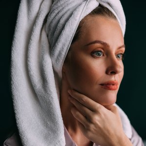 A smiling Caucasian female with her hair wrapped up in a towel keeping her face clean and hydrated using different cosmetic products. (skincare concept)
