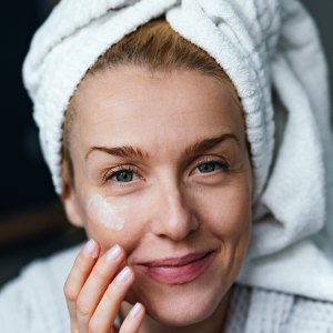 Close up photo of happy woman in bathrobe using face cream and looking at camera while sitting in the bedroom.