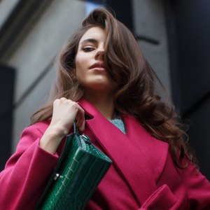 Fabulous portrait of the young stylish brunette. Elegant young woman with long wavy dark hair looks down thoughtfully. She wears stylish crimson coat and green leather bag.