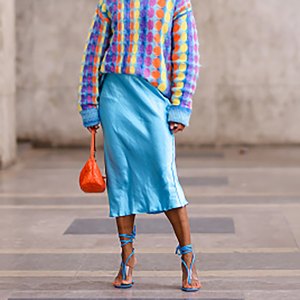 PARIS, FRANCE - APRIL 02: Ellie Delphine wears gold earrings, a gold chain pendant necklace, a pale blue with yellow / orange / purple circle print pattern wool pullover, a pale blue satin midi skirt, a neon orange shiny leather varnished handbag from Miu Miu, blue leather strappy / laces ankle heels flip-flop sandals / shoes , during a street style fashion photo session, on April 02, 2022 in Paris, France. (Photo by Edward Berthelot/Getty Images)