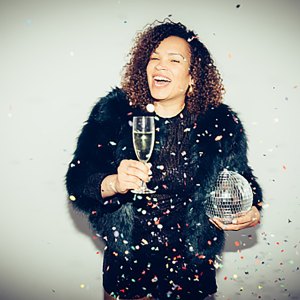A smiling woman is holding a glass of champagne and a disco ball, wearing a black party dress amid falling confetti against a spot lit white background