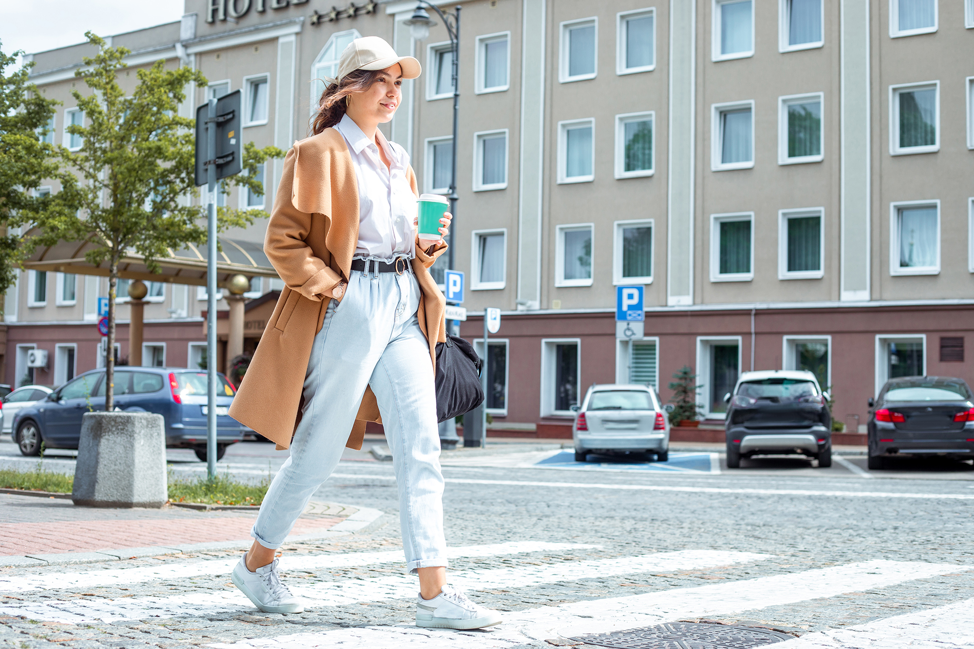 Afternoon stroll: Confident young lady with casual clothing style strolls along sunlit paved sidewalk in downtown, taking sips from coffee cup, crossing road and embracing urban charm around and nice weather