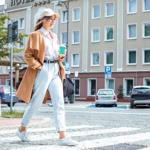 Afternoon stroll: Confident young lady with casual clothing style strolls along sunlit paved sidewalk in downtown, taking sips from coffee cup, crossing road and embracing urban charm around and nice weather