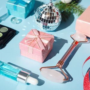 Pink Christmas gift box, rose quartz Jade face roller, cream, face serum, silver Disco balls, fir tree branch, trendy 2022 color eyeshadow, on pastel blue color background. Shiny glittering template for beauty product or cocktail party invitation. Holiday party, Merry Christmas, Happy New Year, Black Friday, Valentine's day and Birthday concept. Minimalistic background with copy space. Close-up. Flat lay.