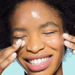 Close-up of woman applying glossy face make-up moisturizer, smiling, with eyes closed with turquoise background
