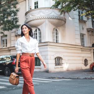 A stylish woman walks confidently down a picturesque street in a charming urban neighborhood, exuding elegance and freedom. She carries a woven handbag and is surrounded by classic architecture and parked cars.