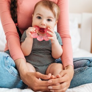 Cute little baby girl playing with teething toy, biting teether and looking at camera, sitting with mother on bed at home. Motherhood and childcare concept