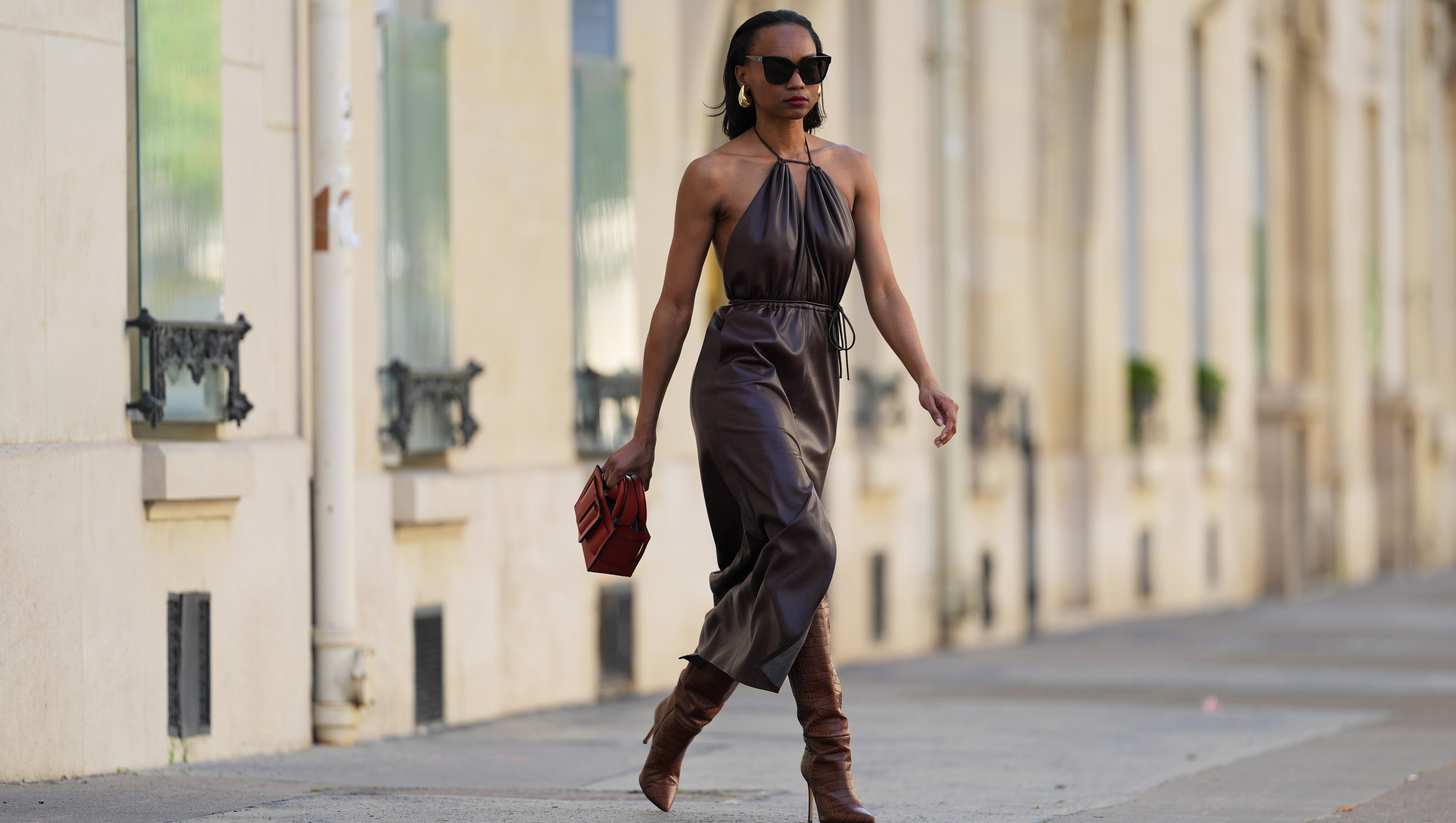 PARIS, FRANCE - APRIL 13: Emilie Joseph wears sunglasses, golden earrings, a brown leather gathered sleeveless dress, a red leather bag, brown leather boots / pointed shoes high heels with crocodile patterns, during a street style fashion photo session, on April 13, 2024 in Paris, France. (Photo by Edward Berthelot/Getty Images)