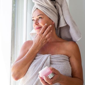 Smiling woman applying moisturizer on face standing by window at home