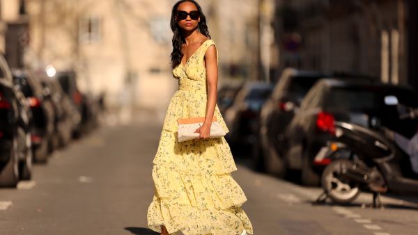 Emilie Joseph @in_fashionwetrust wears black sunglasses, a pale yellow with green and yellow flower print pattern V-neck / tank-top / backless / belted / ruffled maxi long dress, a beige fabric ruffled clutch, silver rings, pale yellow matte leather pointed pumps heels shoes, during a street style fashion photo session, on March 26, 2022 in Paris, France.
