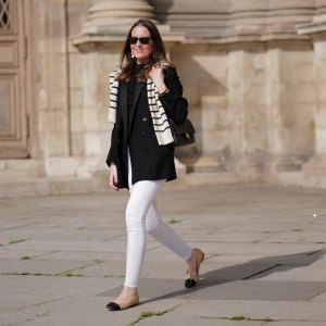 Alba Garavito Torre wears black sunglasses from Ray Ban, gold and white pendant large earrings, a black t-shirt, a white with black small striped print pattern wool pullover from Massimo Dutti, a black shiny grained leather Timeless shoulder bag from Chanel, a black oversized blazer jacket from Munthe, white skinny denim pants from Zara, beige shiny leather with black toe-cap ballerinas from Chanel , during a street style fashion photo session, on April 29, 2023 in Paris, France.