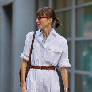 NEW YORK, NEW YORK - SEPTEMBER 15: A guest wears their hair pulled back into a low ponytail with straight bangs, small round burgundy sunglasses with dark lenses, a silver metal watch with a black face on the left wrist, a brown leather shoulder bag with a thin strap, a white button-front jumpsuit in a lightweight woven fabric with chest flap pockets and rolled sleeves, a blue-and-white striped shirt collar peeking at the neckline, a braided brown leather belt with a gold-toned buckle cinching the waist, outside Zankov, during New York Fashion Week, on September 15, 2025 in New York, New York (Photo by Edward Berthelot/Getty Images)