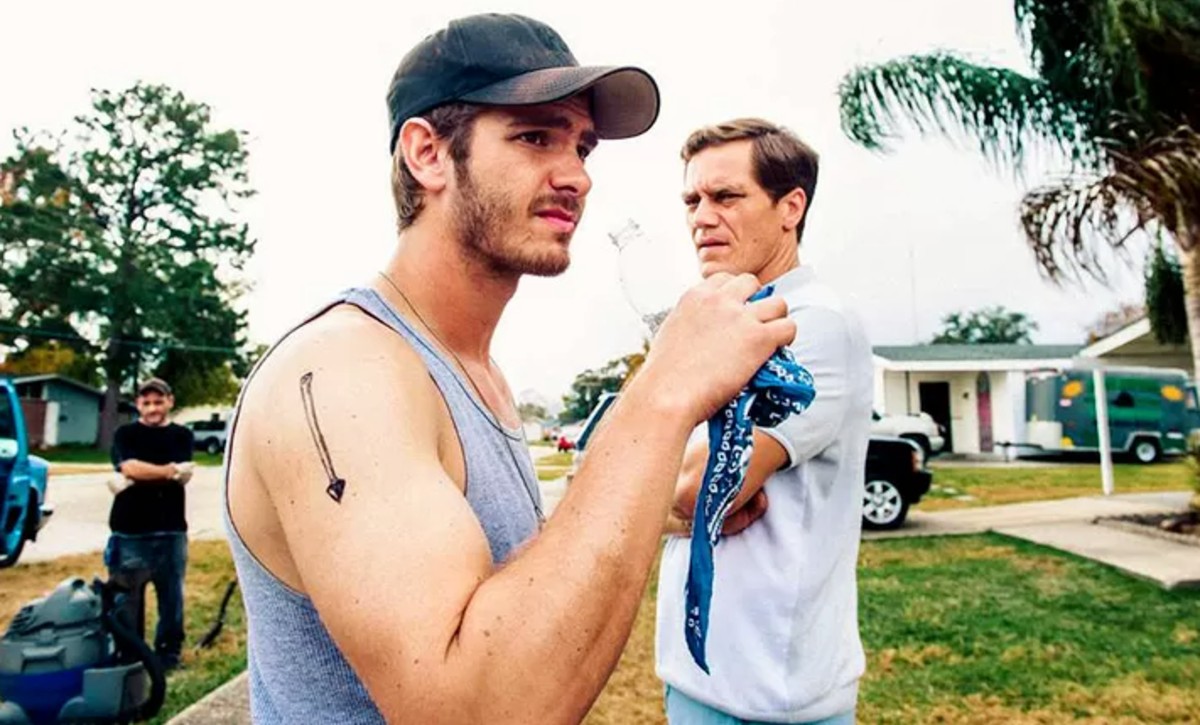 A man drinks water while another man stands behind him in 99 Homes.