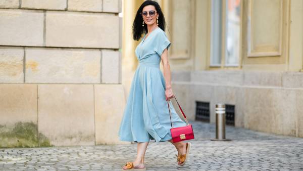 Cosmina Pasarin wears sunglasses, a pastel blue gathered low-neck midi dress, a red bag, brown sandals with platform beige wicker platforms, on July 27, 2021 in Sibiu, Romania.