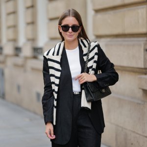 Diane Batoukina wears black large sunglasses, a white halter-neck tank-top from Anine Bing, a black blazer jacket from Massimo Dutti, a white latte and black striped print pattern wool pullover from Zara, a black shiny grained leather small Kelly belt from Hermes, black suit shorts from Massimo Dutti, a black shiny leather quilted Timeless handbag from Chanel, during a street style fashion photo session, on April 28, 2023 in Paris, France.