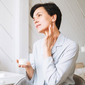 Young adult brunette woman with short hair applies cream to her face at home, morning skincare routine - stock photo
