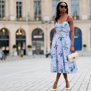 PARIS, FRANCE - APRIL 29: Emilie Joseph wears black large sunglasses, crystal earrings, a pale blue with red / green / navy blue / beige flower print pattern heart neck / tank-top / belted / midi dress, gold bracelets, a beige wickers and white shiny leather bi-material handbag, yellow shiny leather pointed pumps heels shoes , during a street style fashion photo session, on April 29, 2023 in Paris, France. (Photo by Edward Berthelot/Getty Images)