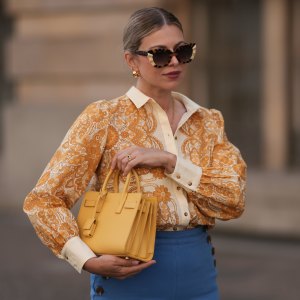 BERLIN, GERMANY - JUNE 07: Aline Kaplan is seen wearing large sunglasses with beige, brown and black pattern from Tibi; chunky golden hoop earrings; several golden necklaces; a creme-white blouse with floral orange pattern from Sézane; a blue high-waisted pair of trousers with two button borders on the hips from Sézane; golden rings and a yellow leather handbag from Saint Laurent on June 07, 2024 in Berlin, Germany. (Photo by Jeremy Moeller/Getty Images)