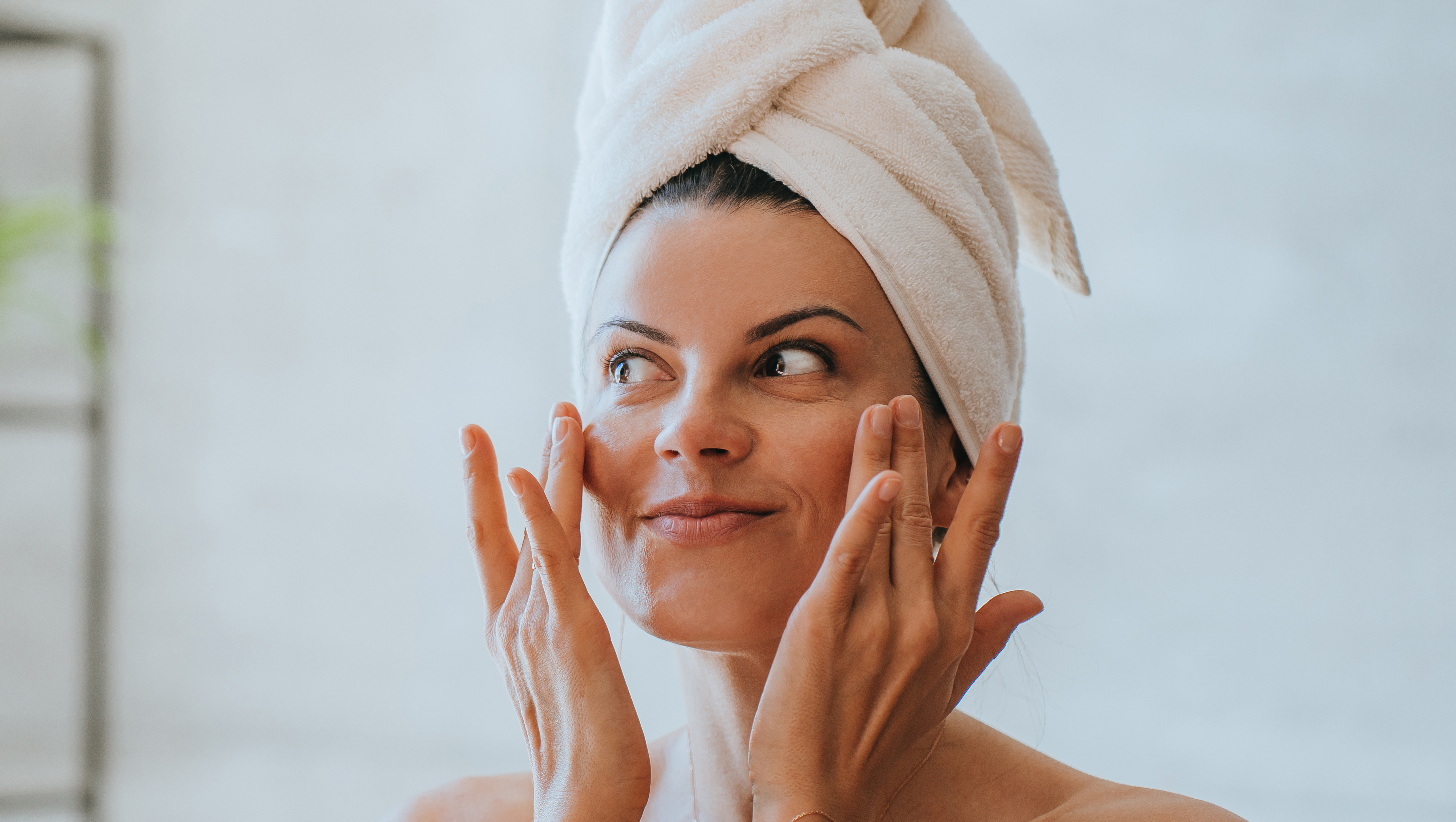 Young adult hispanic woman at bathroom with towel wrapped around head applying cream on face satisfied by spa procedure. Relaxed American female trying new skin care lotion at hotel. Healthy life.