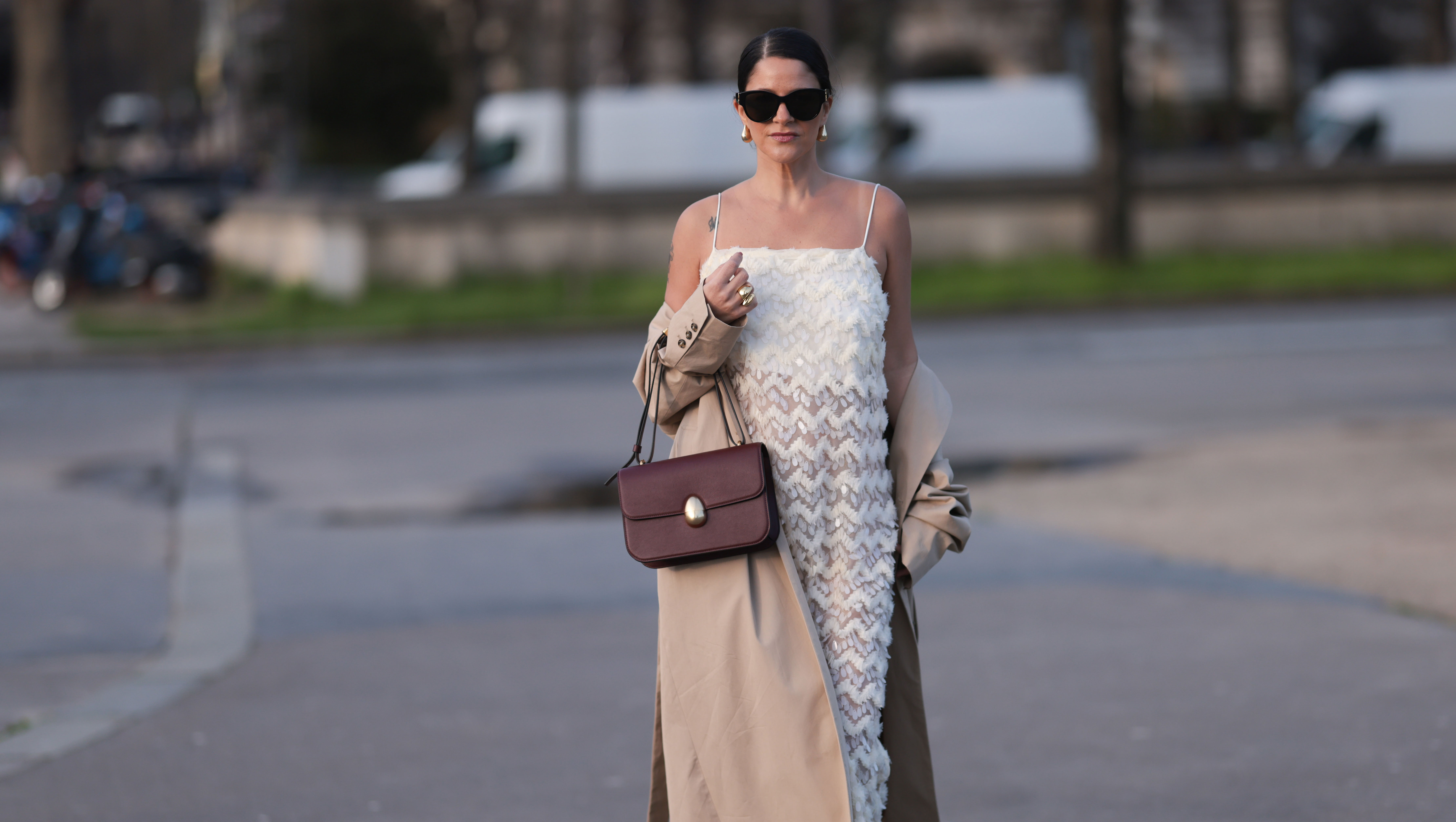 PARIS, FRANCE - FEBRUARY 27: Gili Biegun seen wearing YSL black sunglasses, Bottega Veneta gold tear drops earrings, white long sheer pearl embroidered maxi dress, gold statement rings, beige oversized long coat, Neous burgundy leather bag and burgundy varnished leather pumps / heels, outside Anrealage, during the Womenswear Fall/Winter 2024/2025 as part of Paris Fashion Week on February 27, 2024 in Paris, France. (Photo by Jeremy Moeller/Getty Images)