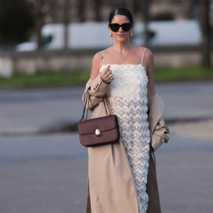 PARIS, FRANCE - FEBRUARY 27: Gili Biegun seen wearing YSL black sunglasses, Bottega Veneta gold tear drops earrings, white long sheer pearl embroidered maxi dress, gold statement rings, beige oversized long coat, Neous burgundy leather bag and burgundy varnished leather pumps / heels, outside Anrealage, during the Womenswear Fall/Winter 2024/2025 as part of Paris Fashion Week on February 27, 2024 in Paris, France. (Photo by Jeremy Moeller/Getty Images)