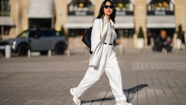 Xiayan wears black Bottega Veneta sunglasses, silver earrings, a silver chain necklace, a white V-neck silk gown, a white long oversized Sundarbay blazer jacket with white suit Sundarbay matching pants, a black shiny leather belt, white and beige with gold details Stellaluna sneakers, a black shiny leather Louis Vuitton monogram-embossed Coussin PM crossbody bag, on February 27, 2021 in Paris, France.