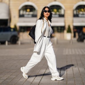 Xiayan wears black Bottega Veneta sunglasses, silver earrings, a silver chain necklace, a white V-neck silk gown, a white long oversized Sundarbay blazer jacket with white suit Sundarbay matching pants, a black shiny leather belt, white and beige with gold details Stellaluna sneakers, a black shiny leather Louis Vuitton monogram-embossed Coussin PM crossbody bag, on February 27, 2021 in Paris, France.