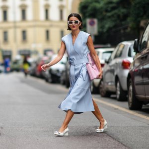 Alexandra Pereira wears white sunglasses, silver and rhinestones earrings, a pale blue shiny leather V-neck / sleeveless / belted / slit midi dress, a pale pink handbag from Salvatore Ferragamo, white leather block heels shoes with embroidered knot on the toe-cap, rings, outside the Ferragamo fashion show during the Milan Fashion Week - Spring / Summer 2022 on September 25, 2021 in Milan, Italy.