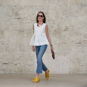Alba Garavito Torre wears black sunglasses, long straight brown hair with a center part, a white pleated peplum sleeveless blouse by Commense, and blue high-rise raw-hem denim jeans. A yellow clog sandal with floral detailing by AGL is worn, paired with a jean denim and brown mini Jackie handbag by Gucci. Accessories include a chunky necklace with colorful natural stones by Sempiterno Paris and gold jewelry. Full body shot, during a street style fashion photo session, on May 02, 2025 in Paris, France.