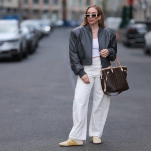 BERLIN, GERMANY - APRIL 09: Sonia Lyson seen wearing Linda Farrow bold white rectangular sunglasses that add a trendy touch, Zara white cotton fitted crop top, Zara high-waisted, wide-leg white relaxed jeans / denim pants, The Frankie Shop black, slightly glossy bomber jacket, Louis Vuitton brown Neverfull tote bag in the classic monogram print and Louis Vuitton beige Sneakerina sneakers / shoes with mustard yellow details, introducing a playful, sporty contrast to the otherwise minimalist outfit, on April 09, 2025 in Berlin, Germany. (Photo by Jeremy Moeller/Getty Images)
