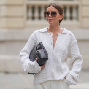 PARIS, FRANCE - APRIL 17: Diane Batoukina wears a white ribbed knit cardigan with a wide collar and oversized sleeves by Lovechild 1979, paired with high-waisted ivory trousers by Herskind. A dark brown leather clutch bag by St. Agni is held under one arm. Oversized square Dior sunglasses with brown gradient lenses are worn. Hair is parted at the center and styled in a sleek low bun. Natural makeup with even skin and soft pink lips is visible. Mid shot, during a street style fashion photo session, on April 17, 2025 in Paris, France. (Photo by Edward Berthelot/Getty Images)