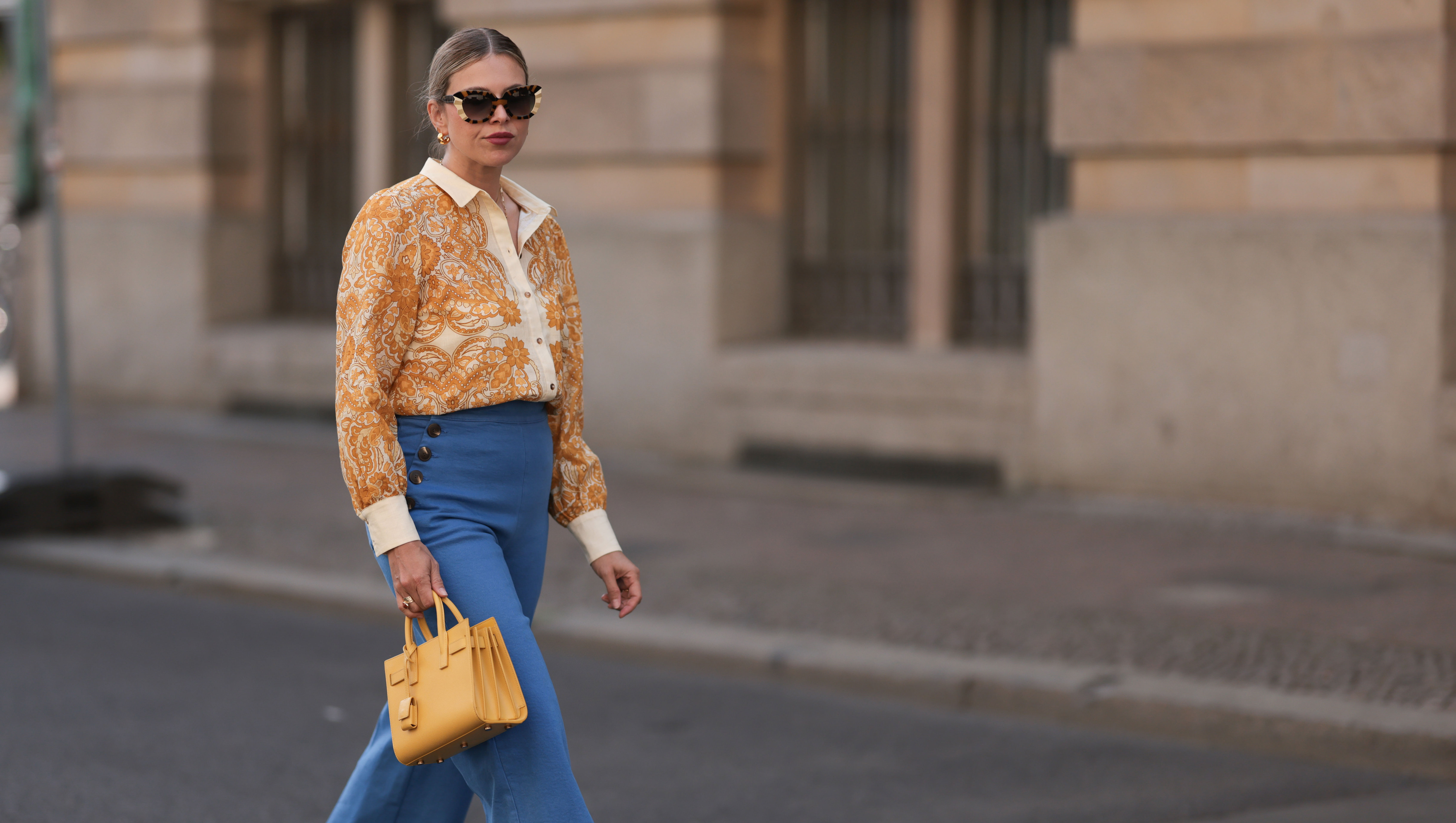 BERLIN, GERMANY - JUNE 07: Aline Kaplan is seen wearing large sunglasses with beige, brown and black pattern from Tibi; chunky golden hoop earrings; several golden necklaces; a creme-white blouse with floral orange pattern from Sézane; a blue high-waisted pair of trousers with two button borders on the hips from Sézane; golden rings; a yellow leather handbag from Saint Laurent and creme-white leather mules from Mango on June 07, 2024 in Berlin, Germany. (Photo by Jeremy Moeller/Getty Images)