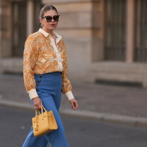 BERLIN, GERMANY - JUNE 07: Aline Kaplan is seen wearing large sunglasses with beige, brown and black pattern from Tibi; chunky golden hoop earrings; several golden necklaces; a creme-white blouse with floral orange pattern from Sézane; a blue high-waisted pair of trousers with two button borders on the hips from Sézane; golden rings; a yellow leather handbag from Saint Laurent and creme-white leather mules from Mango on June 07, 2024 in Berlin, Germany. (Photo by Jeremy Moeller/Getty Images)