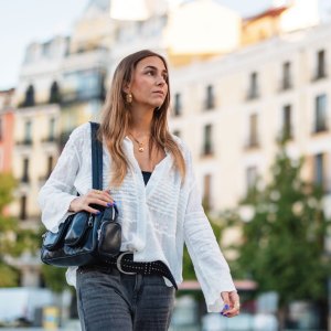 A Hispanic woman walks through a scenic street in Madrid. The background shows traditional Spanish buildings. She carries a handbag and enjoys the urban environment, showcasing everyday life and travel enjoyment.